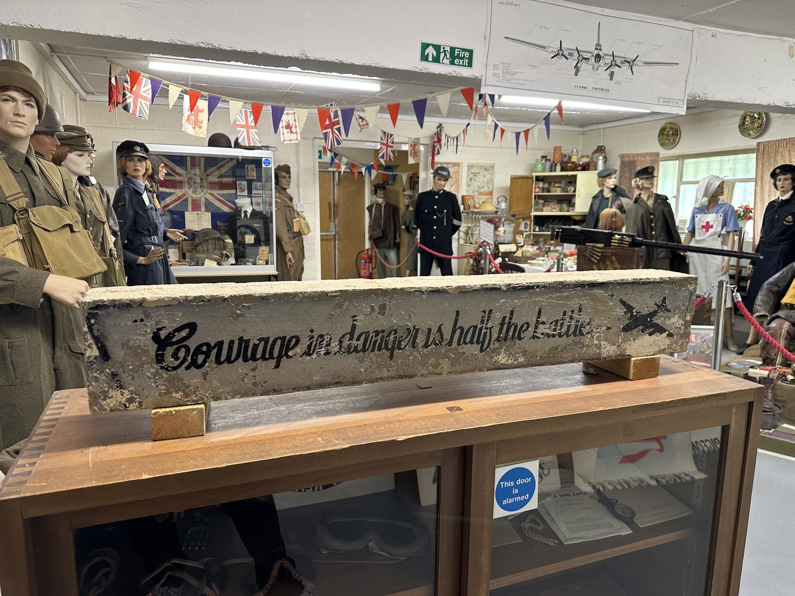 A weathered concrete beam painted with the words Courage in danger is half the battle, mounted on a display case in front of uniformed mannequins and wartime bunting.