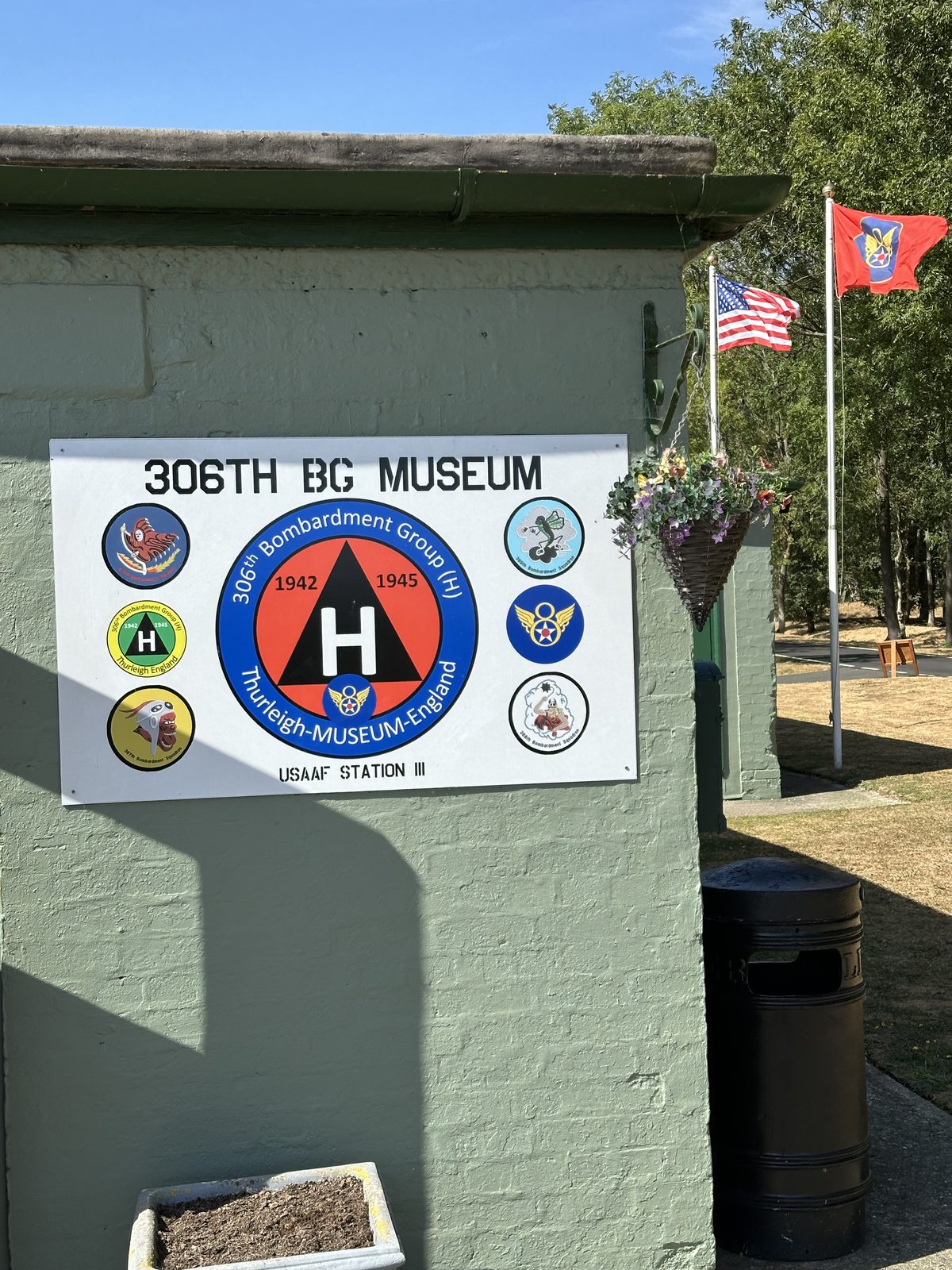 A painted sign reading 306TH BG MUSEUM on the green brick exterior of the museum, carrying the group and squadron insignia and the words USAAF Station 111, with the Stars and Stripes and a squadron flag flying beside it.