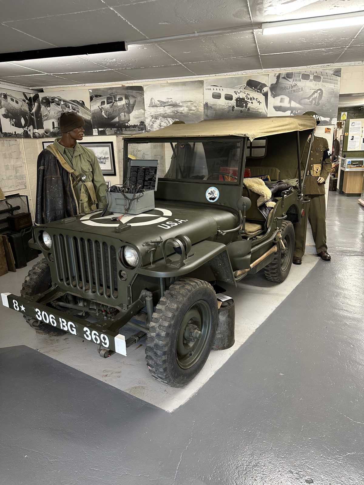 A restored olive-drab Jeep marked U.S.A. with a white star, its bumper stencilled 8 star 306 BG 369, parked below a wall of B-17 nose-art photographs.
