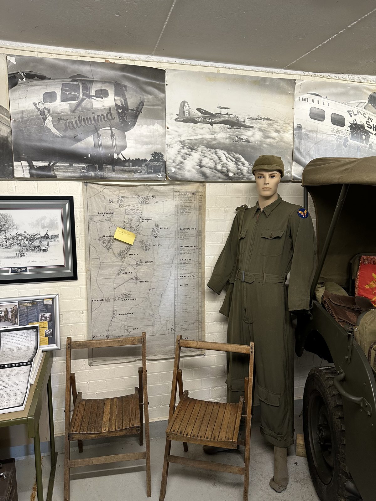Large B-17 nose-art photographs, one lettered Tailwind, hung above a wartime map of the airfield, with a mannequin in a flying coverall and two wooden folding chairs below.