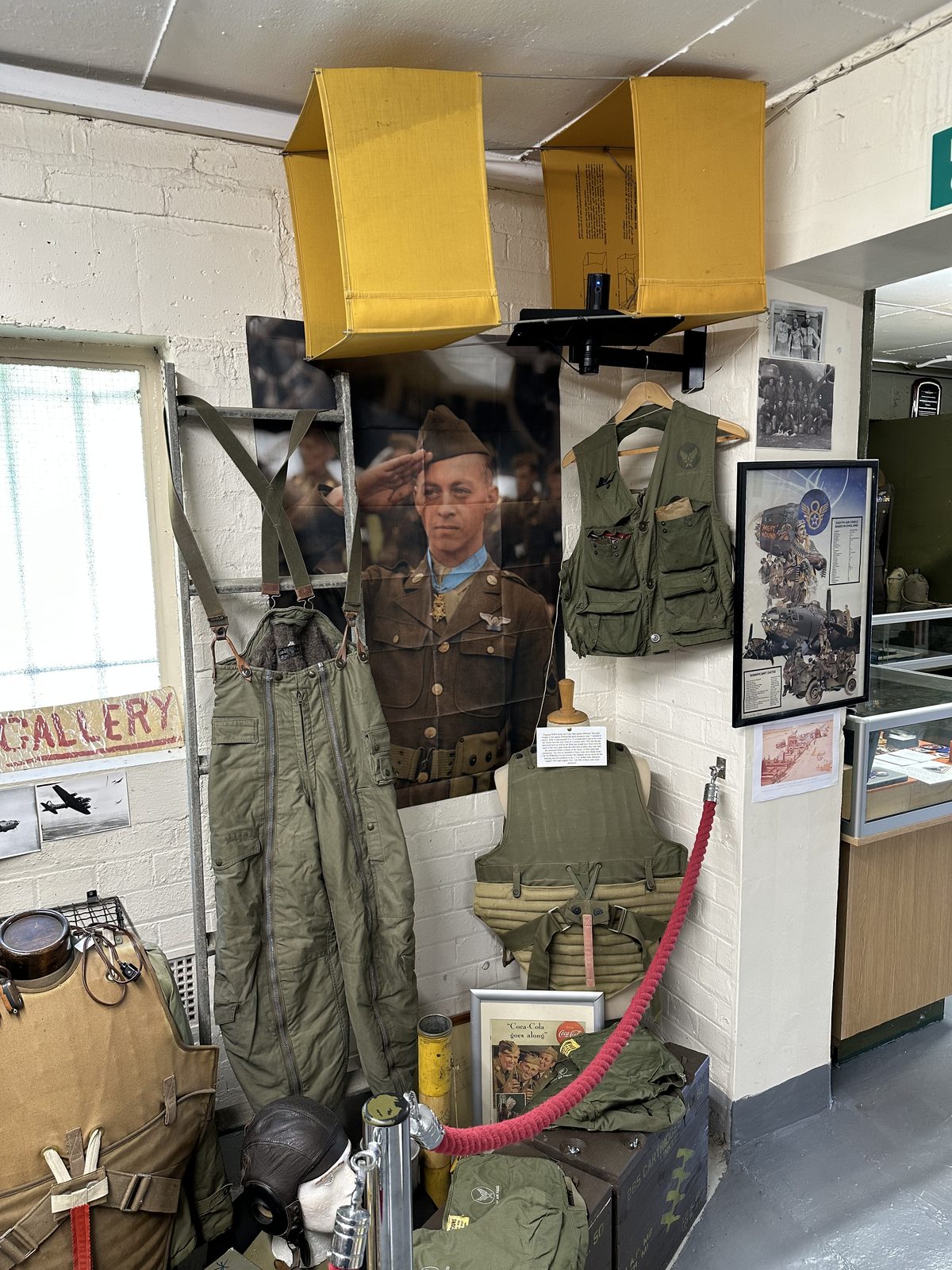 A corner display of flying clothing: heated trousers and a flak vest hung on a rail, a leather helmet and oxygen mask below, under yellow ammunition chutes and a large photograph of a saluting airman wearing the Medal of Honor.
