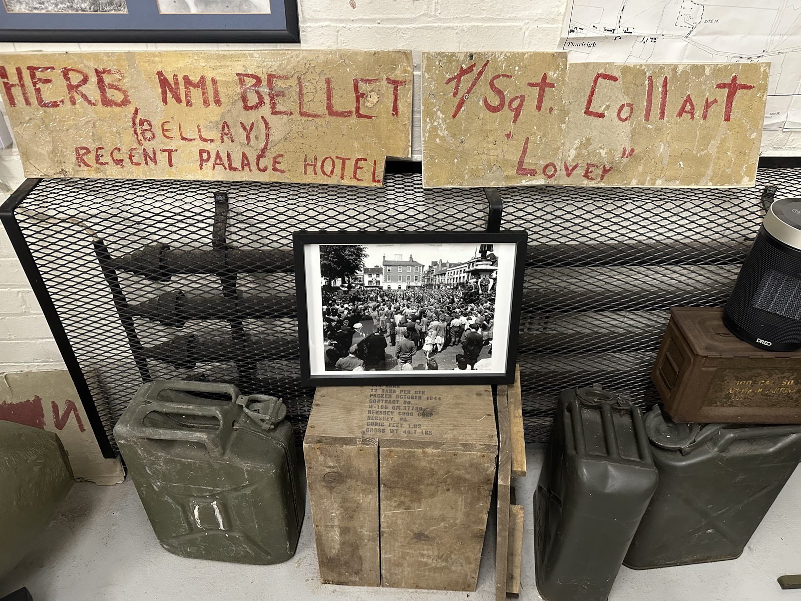 Two salvaged boards hand-painted in red with the names Herb NMI Bellet, Regent Palace Hotel and T/Sgt. Collart, Lover, standing above jerrycans and a framed photograph of a wartime crowd.