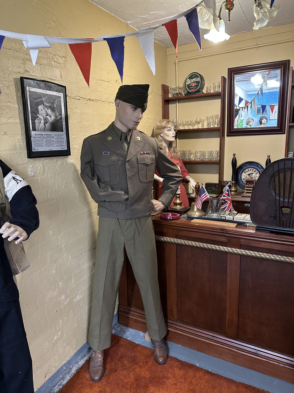 A recreated English pub corner with a bar, glassware, beer pumps and a period radio under red, white and blue bunting, a mannequin in service dress standing beside a figure behind the bar.