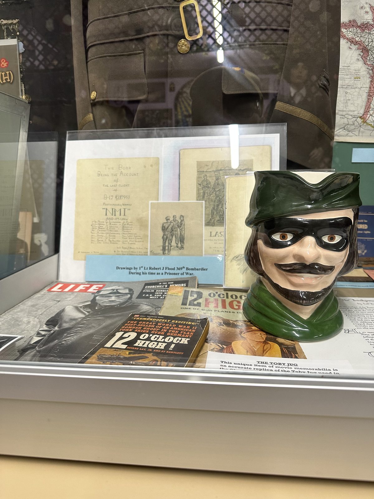 A display case holding a green Toby jug replica from the film Twelve O'Clock High, shown with copies of the novel, a LIFE magazine and a drawing made by Lt. Robert J. Flood while a prisoner of war.