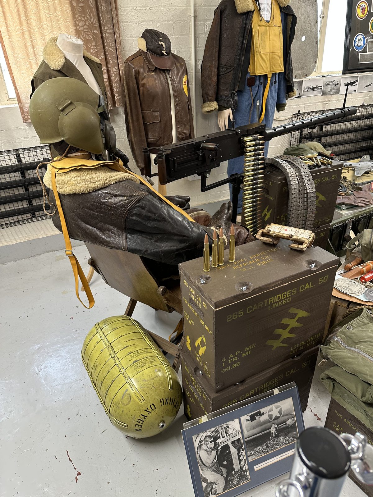 A seated mannequin in a fleece-lined flying jacket at a .50 calibre waist gun, with belted ammunition, stacked cartridge boxes and a yellow oxygen cylinder beside him.